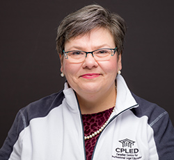 A white women with short grey hair, wearing a white pearl necklace and a white branded CPLED sweater